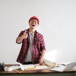 Cheerful male architect in a red hardhat and plaid shirt giving thumbs up at his desk.